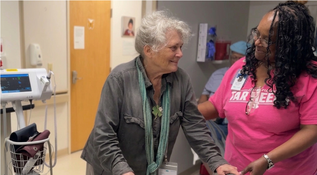 An older woman in a gray shirt smiles at a nurse wearing a pink shirt in a hospital hallway.