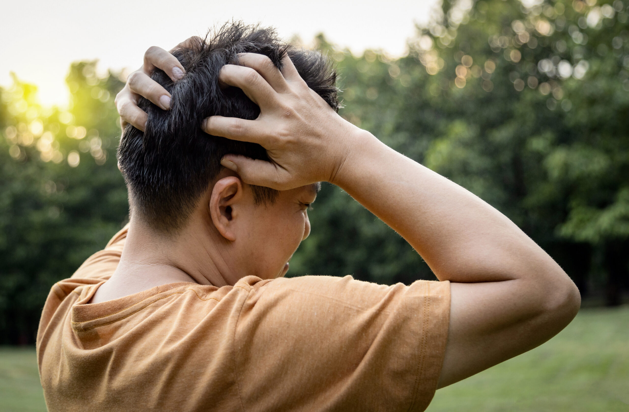 Person standing outdoors with hands clasped on top of their head, back view in a park setting